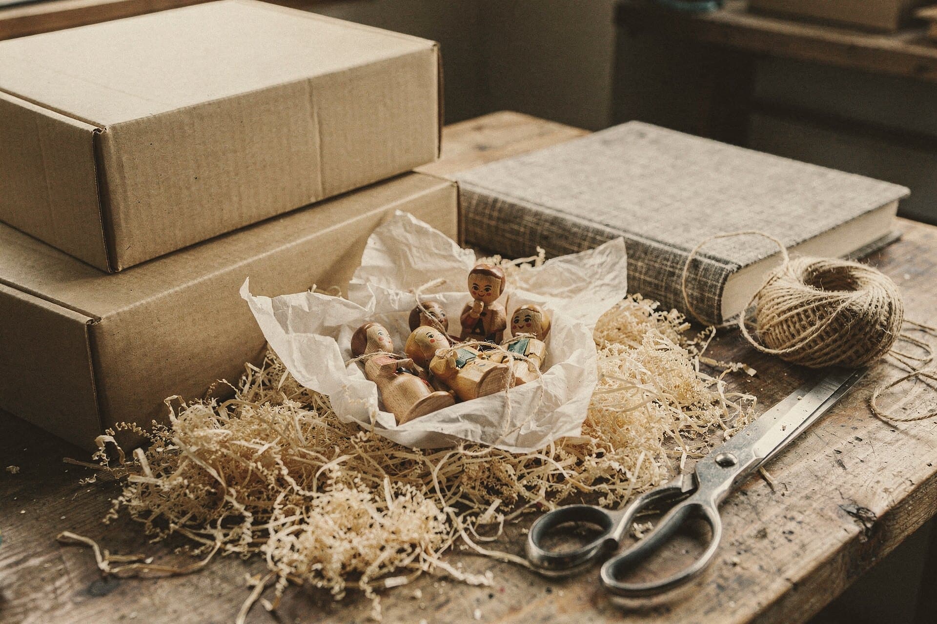 A packing table: figures wrapped in tissue paper on wood wool, plain boxes, a ball of twine, scissors and a clothbound book.