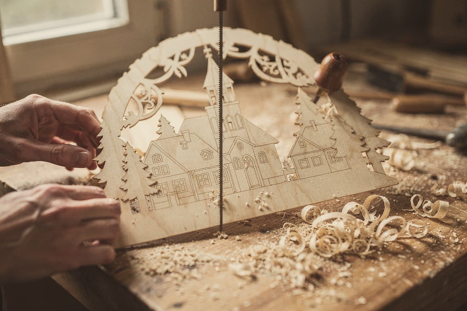 A fretsaw blade cuts the silhouette of a candle arch, a village with church and spruce trees, from thin plywood.