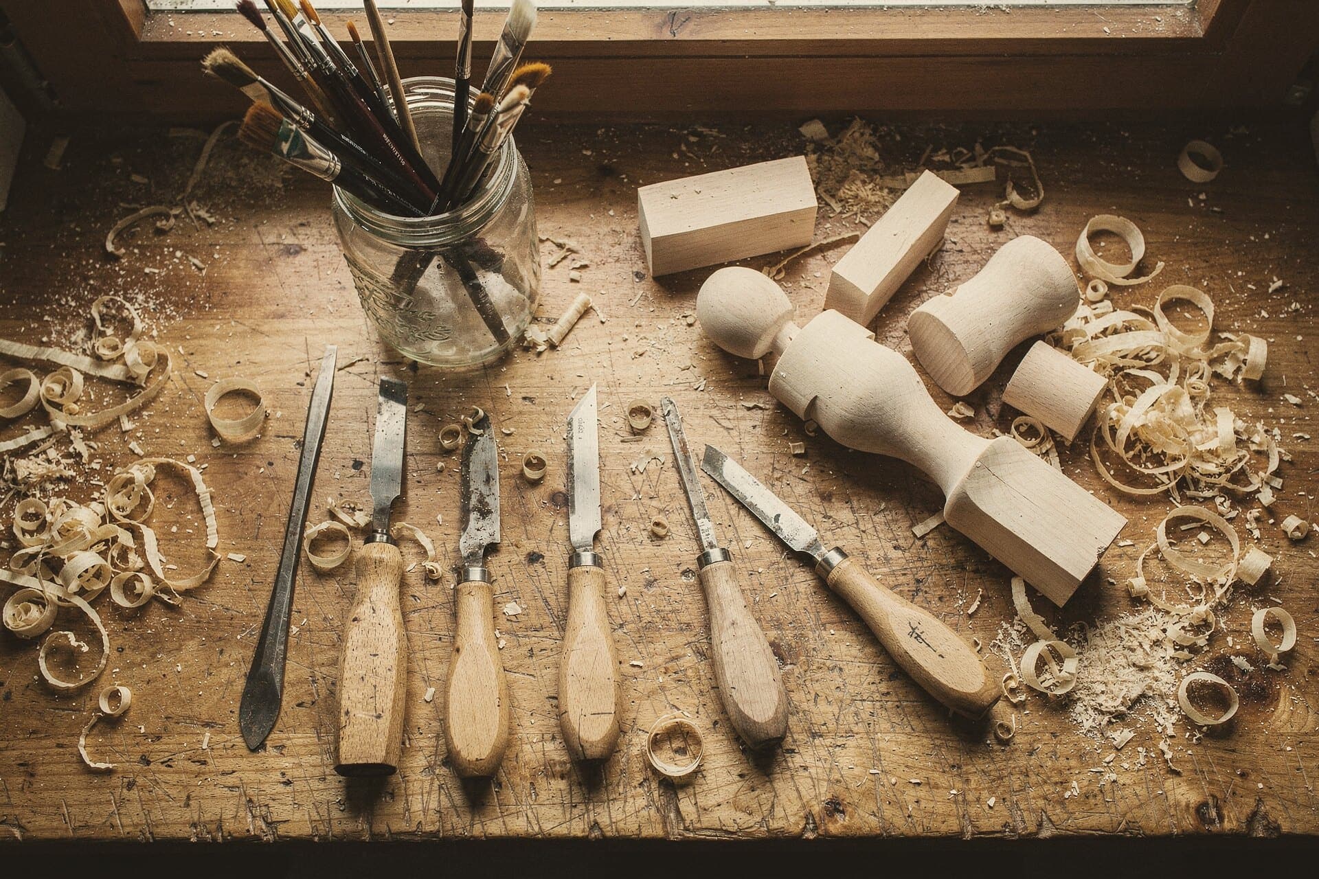 From above: turning chisels and carving knives laid out on a worn bench among linden blanks, a half-turned figure and curls of shavings.