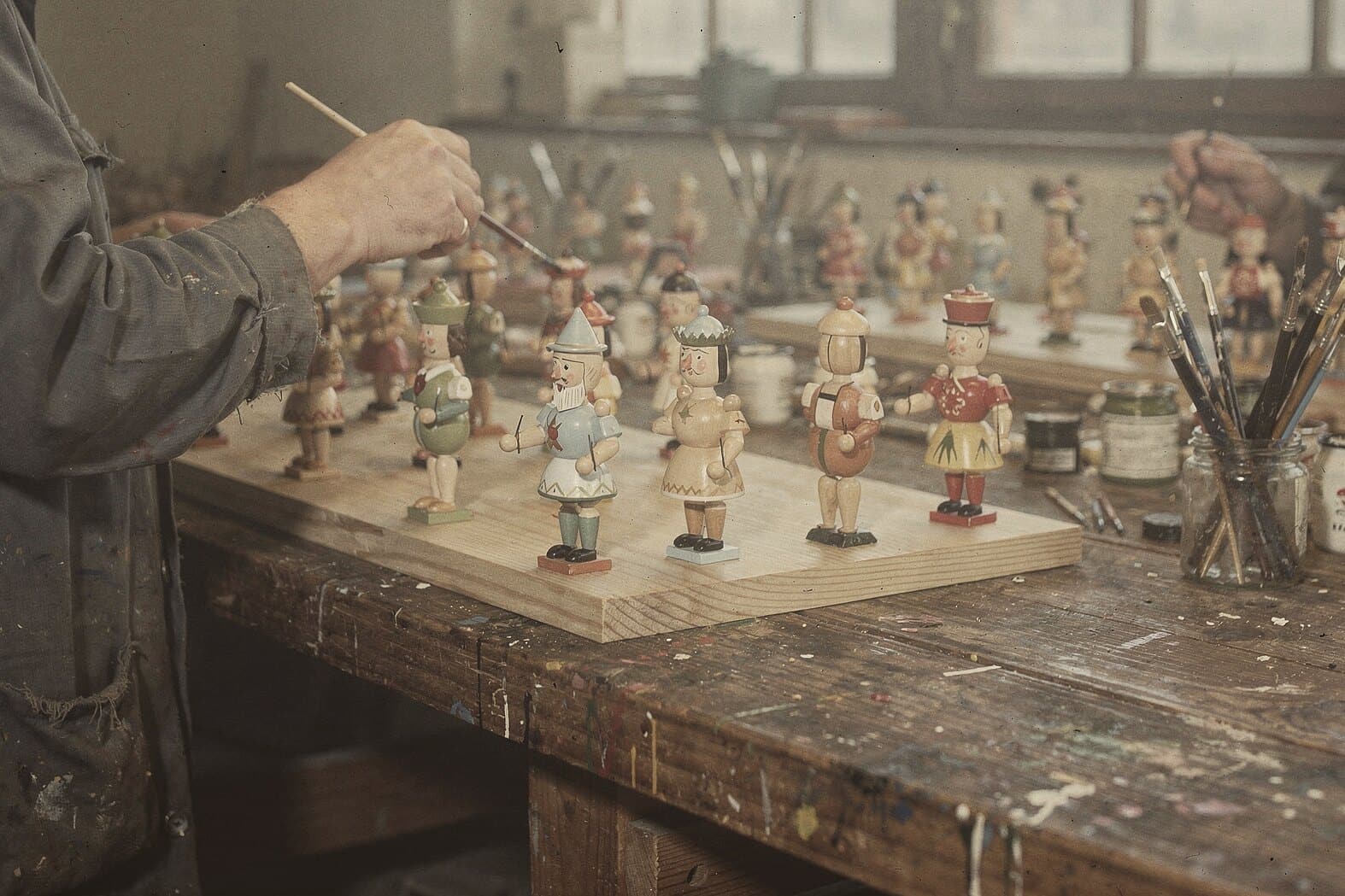 Hands painting rows of small turned figures at a long, paint-spattered workshop table, in faded mid-century color.