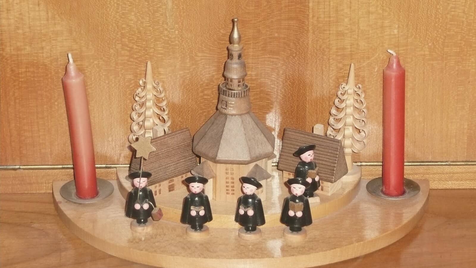 A wooden miniature of Seiffen's church with carol singers in black capes, two red candles and small shaved trees.