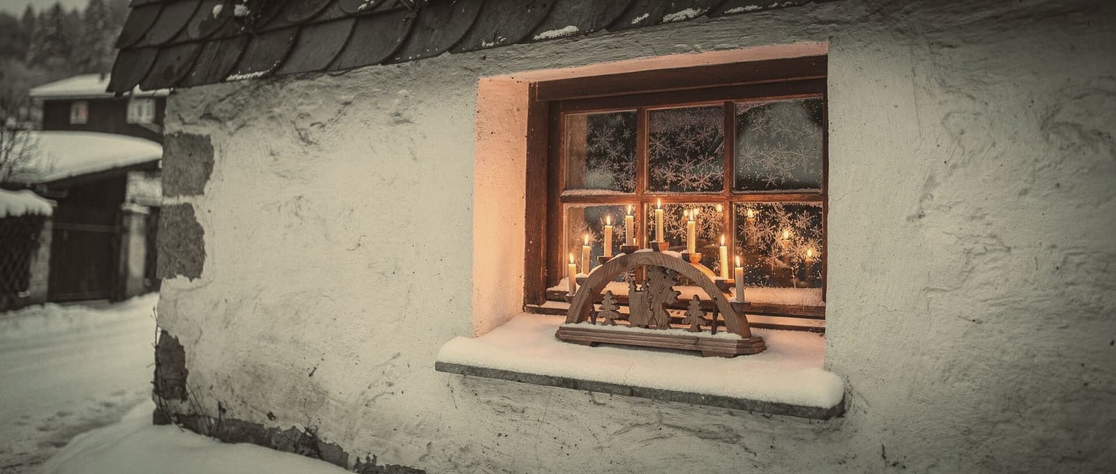 A candle arch glowing in a frosted cottage window at dusk, snow on the sill and on the street outside.
