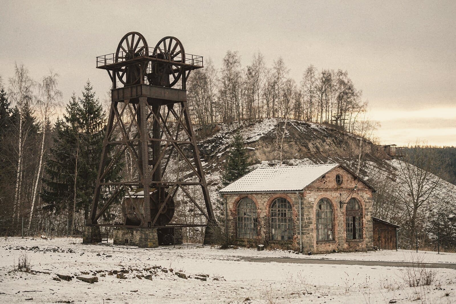 Ein genietetes eisernes Fördergerüst mit zwei Seilscheiben über einem alten Schacht, daneben ein steinernes Maschinenhaus mit Rundbogenfenstern, leichter Schnee.
