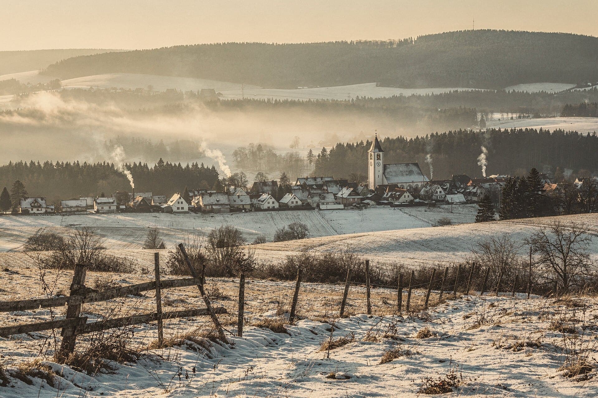 Winter im Erzgebirge: verschneite Felder und Fichtenwald, ein Dorf mit weißem Kirchturm, Nebel im Tal.