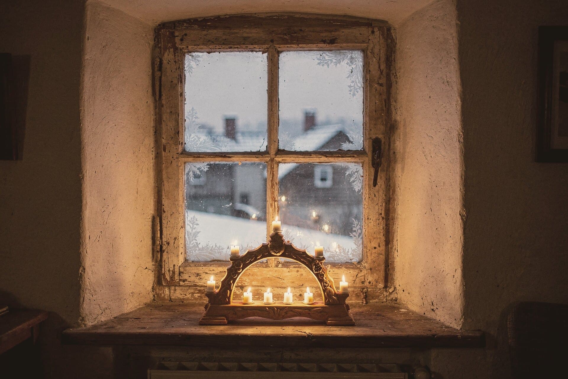 A lit candle arch in a deep window recess at dusk, frost on the panes and the snowy village outside.