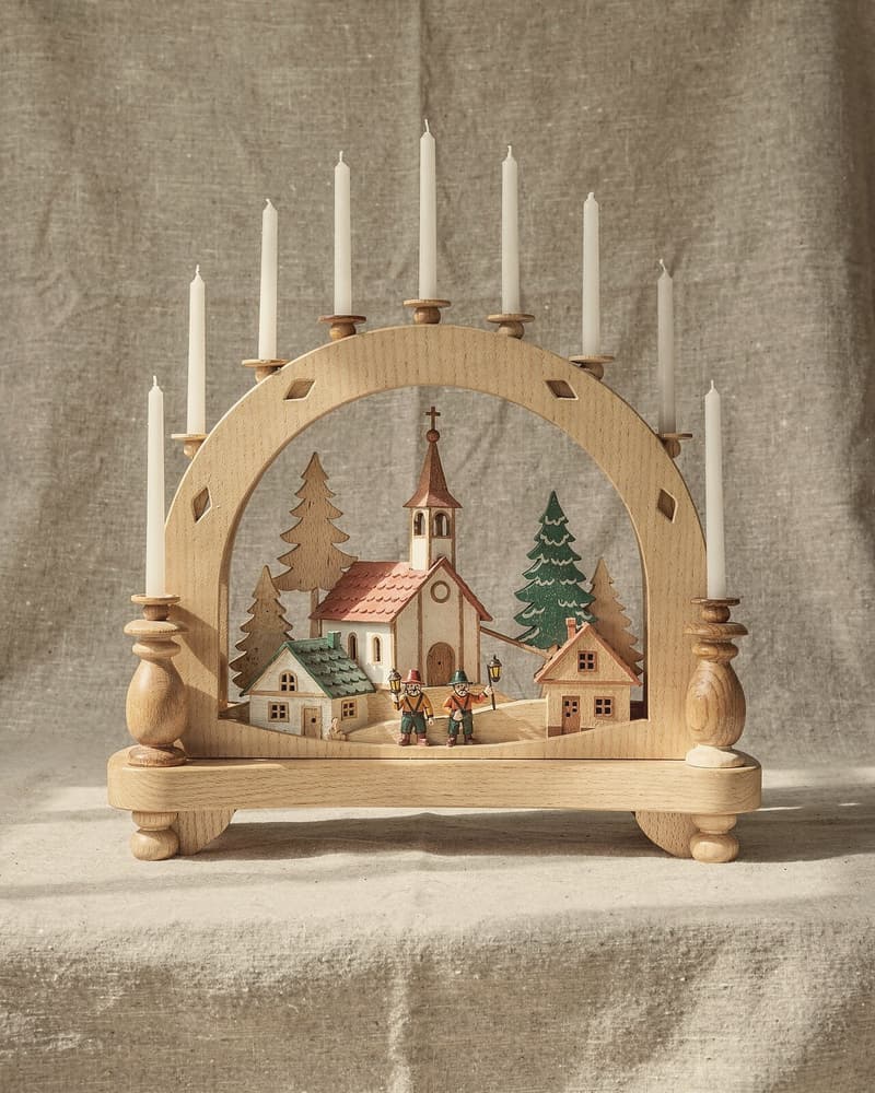 Ein heller Schwibbogen aus Holz mit neun weißen Kerzen; darunter ein ausgesägtes Dorf mit Kirche, Fichten und zwei kleinen Bergleuten.
