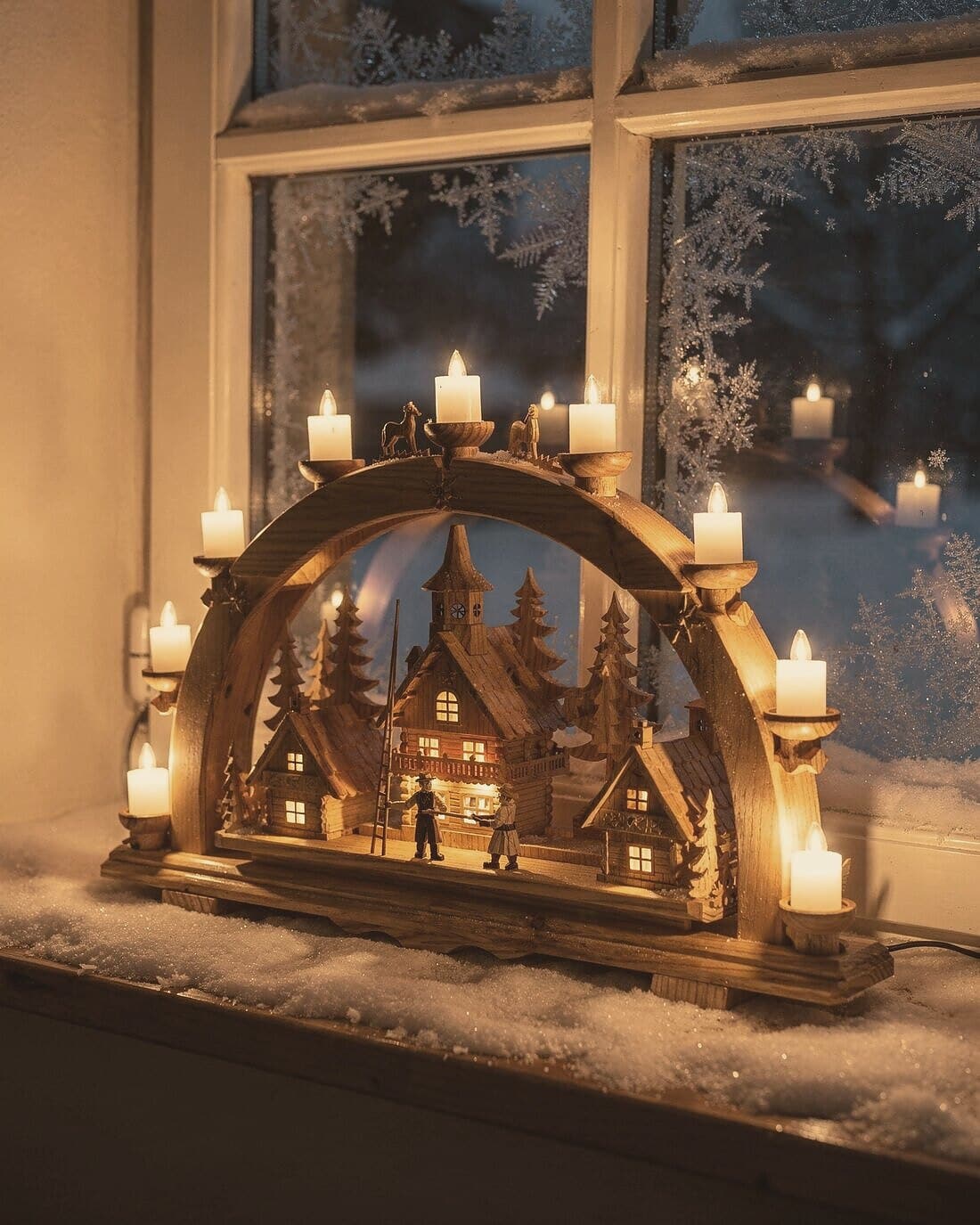 Ein brennender Schwibbogen mit Dorfmotiv im bereiften Fenster, Schnee auf dem Sims.