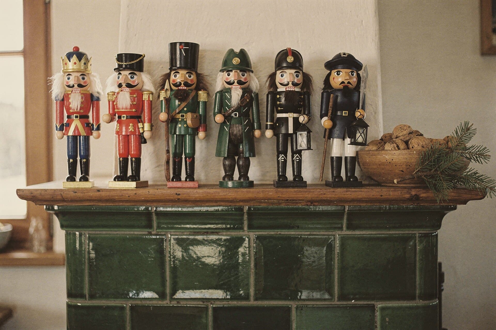 Six nutcrackers in a row on a green tiled stove, a bowl of walnuts and a spruce sprig at the end.