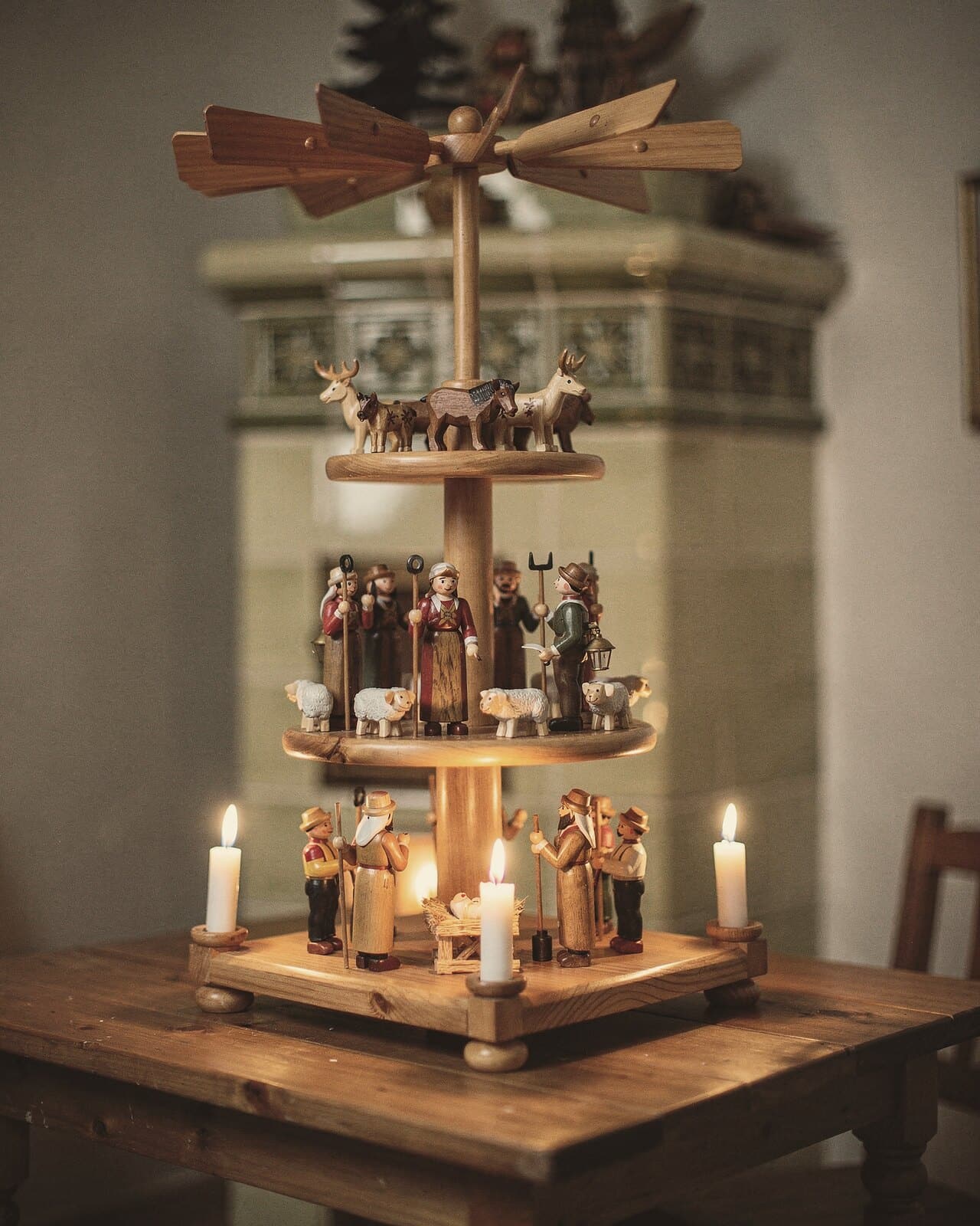Eine hohe mehrstöckige Pyramide mit Krippe, Hirten und Tieren, die Kerzen brennen, dahinter ein Kachelofen.