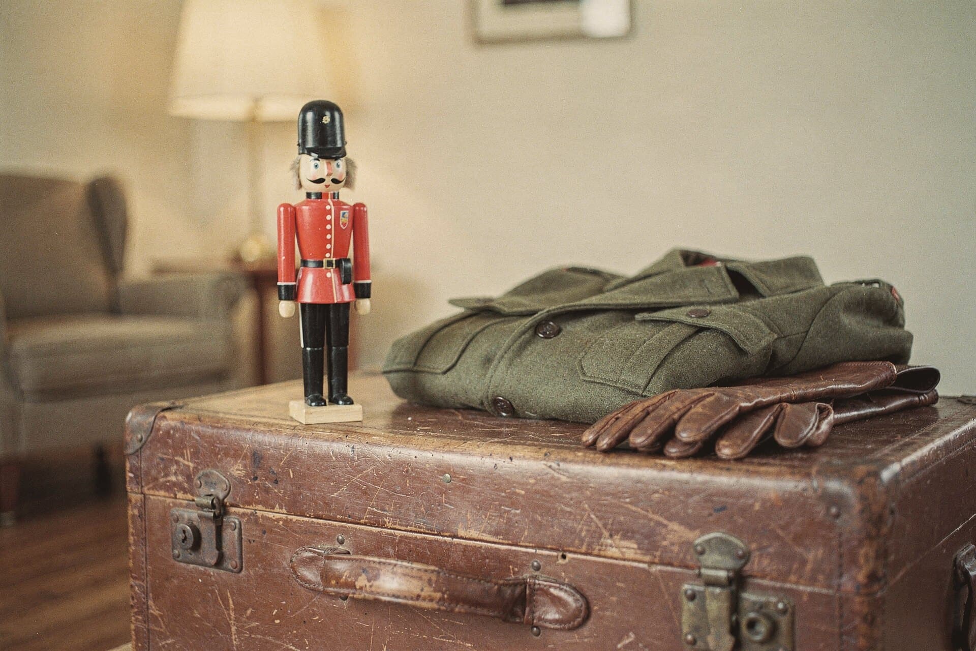 A soldier nutcracker stands on an old leather trunk beside a folded olive wool army coat and leather gloves.