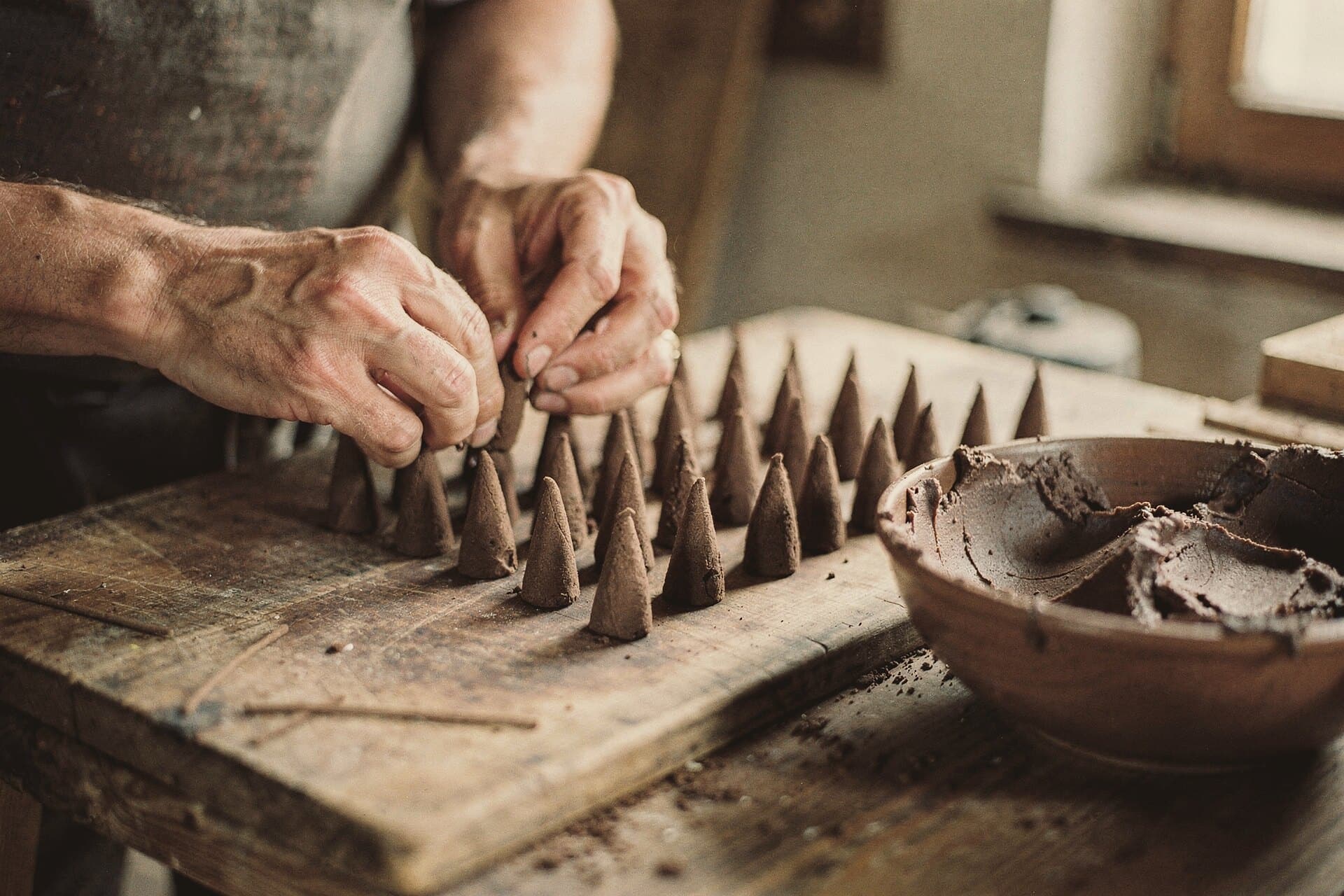 Hands shape incense cones from dark paste on a wooden board, rows of finished cones drying beside a bowl.