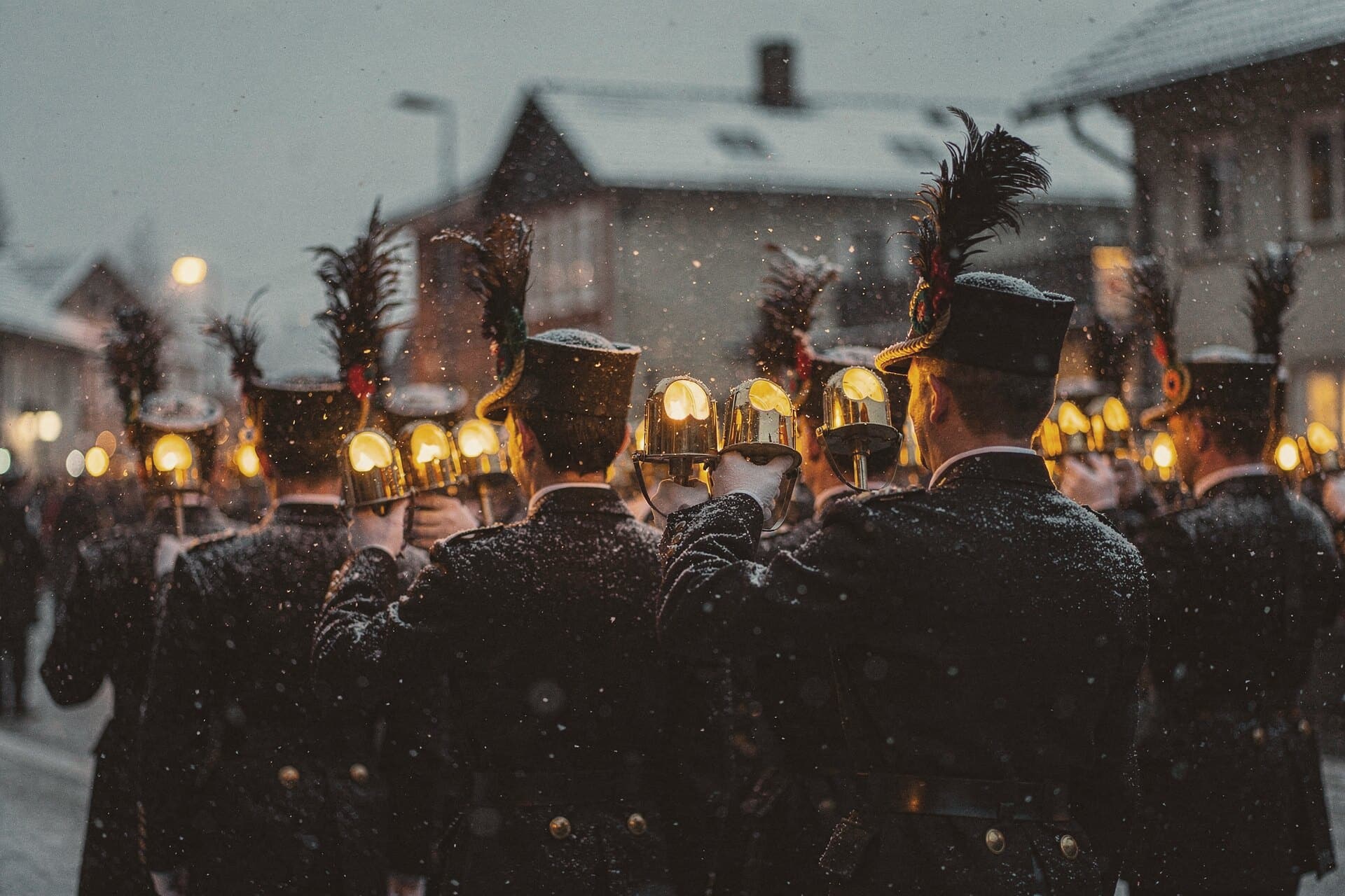 Miners' lamps held high in a parade at dusk, plumed hats and black uniforms seen from behind, snow falling.