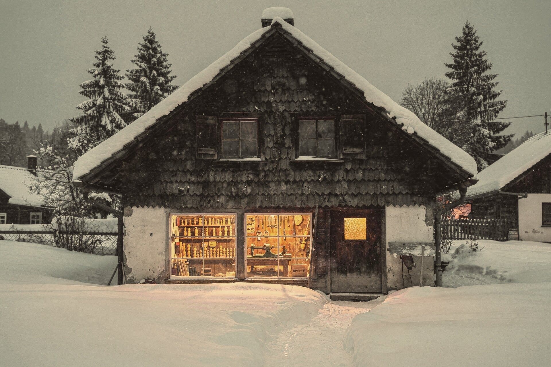 Ein schindelverkleidetes Haus im tiefen Schnee in der Dämmerung, die Werkstattfenster leuchten, drinnen Regale mit Holzfiguren und eine Drehbank.