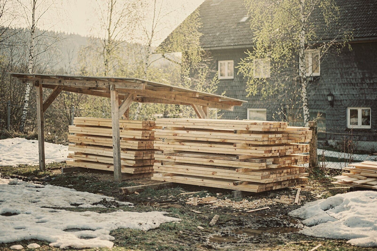 Spring: boards stacked to air-dry under a small roof in the yard, the last snow melting, birches in bud.
